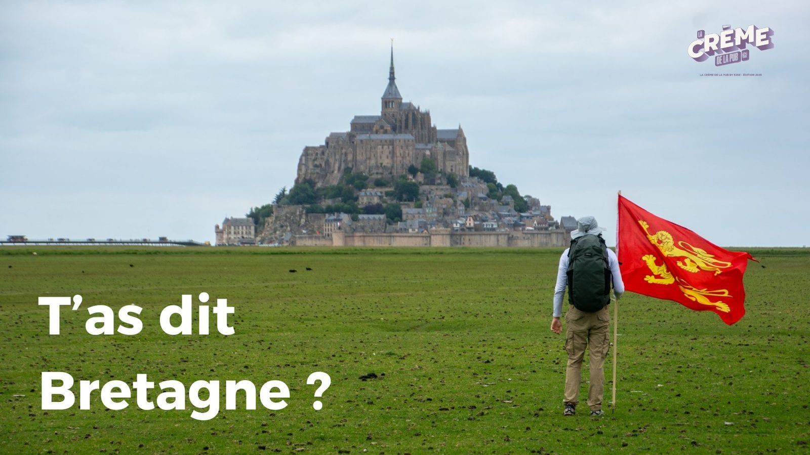 Le Mont-Saint-Michel au loin sous un ciel gris ; au premier plan, un marcheur de dos traverse un pré en portant un grand drapeau normand.