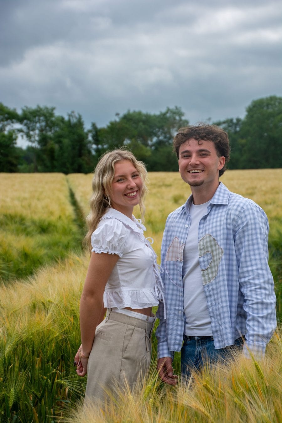 Un couple debout au milieu d’un champ de blé vert, souriant à l’objectif, lisière d’arbres et ciel nuageux derrière eux.