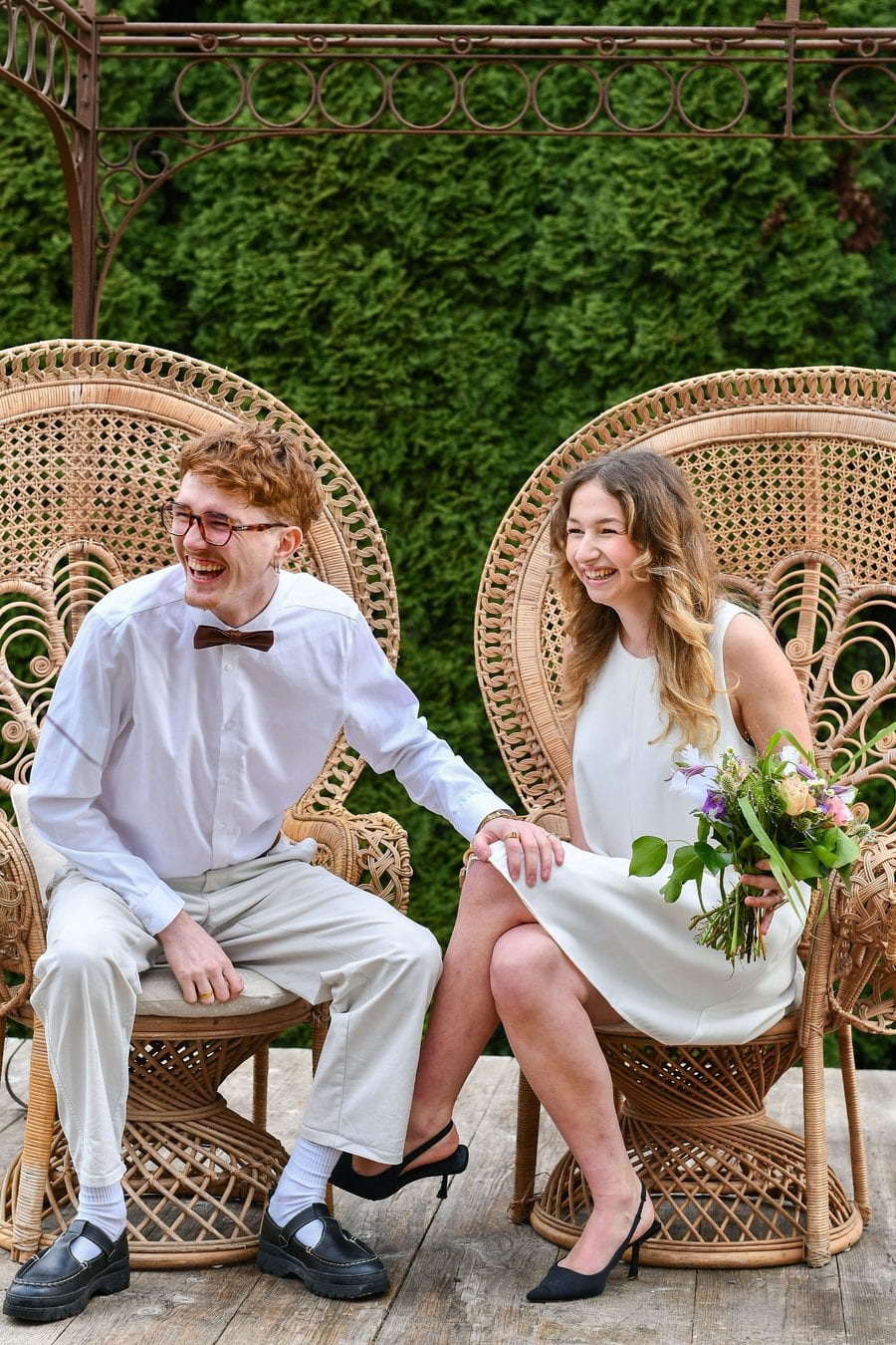 Les mariés assis côte à côte dans deux grands fauteuils en rotin devant un mur de verdure, riant ; elle tient un bouquet.