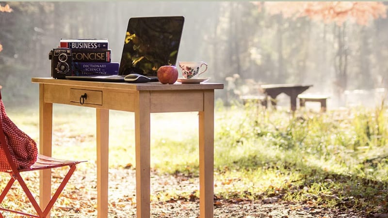Une table en bois dressée en pleine forêt au lever du jour, avec un ordinateur portable, une pile de livres, une tasse et une pomme ; une chaise pliante à côté.