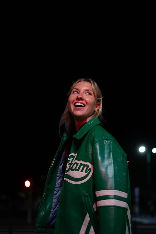 Portrait de nuit d’une jeune femme en veste de baseball verte, riant, le regard levé, devant les lumières floues de la ville.